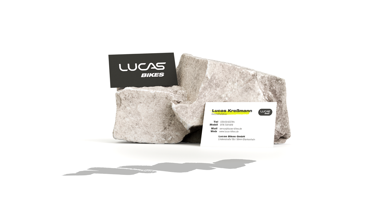 Zwei Visitenkarten, eine schwarze mit dem Logo von Lucas Bikes und eine weiße mit den Kontaktdaten, stehen auf rauen, hellen Steinen - ähnlich wie bei der Tanzschule Fischer - und werfen Schatten auf einen sauberen, weißen Hintergrund.