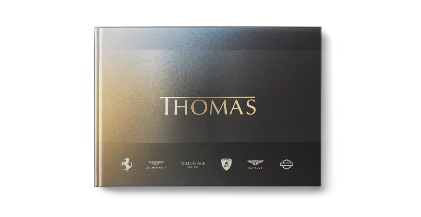 Ein gebundenes Buch mit einem schwarzen Einband mit Farbverlauf, dem Namen „THOMAS“ in goldenen Buchstaben und Logos von Luxusautomarken (Ferrari, Aston Martin, Rolls-Royce, Lamborghini, Bentley, Harley-Davidson) am unteren Rand.